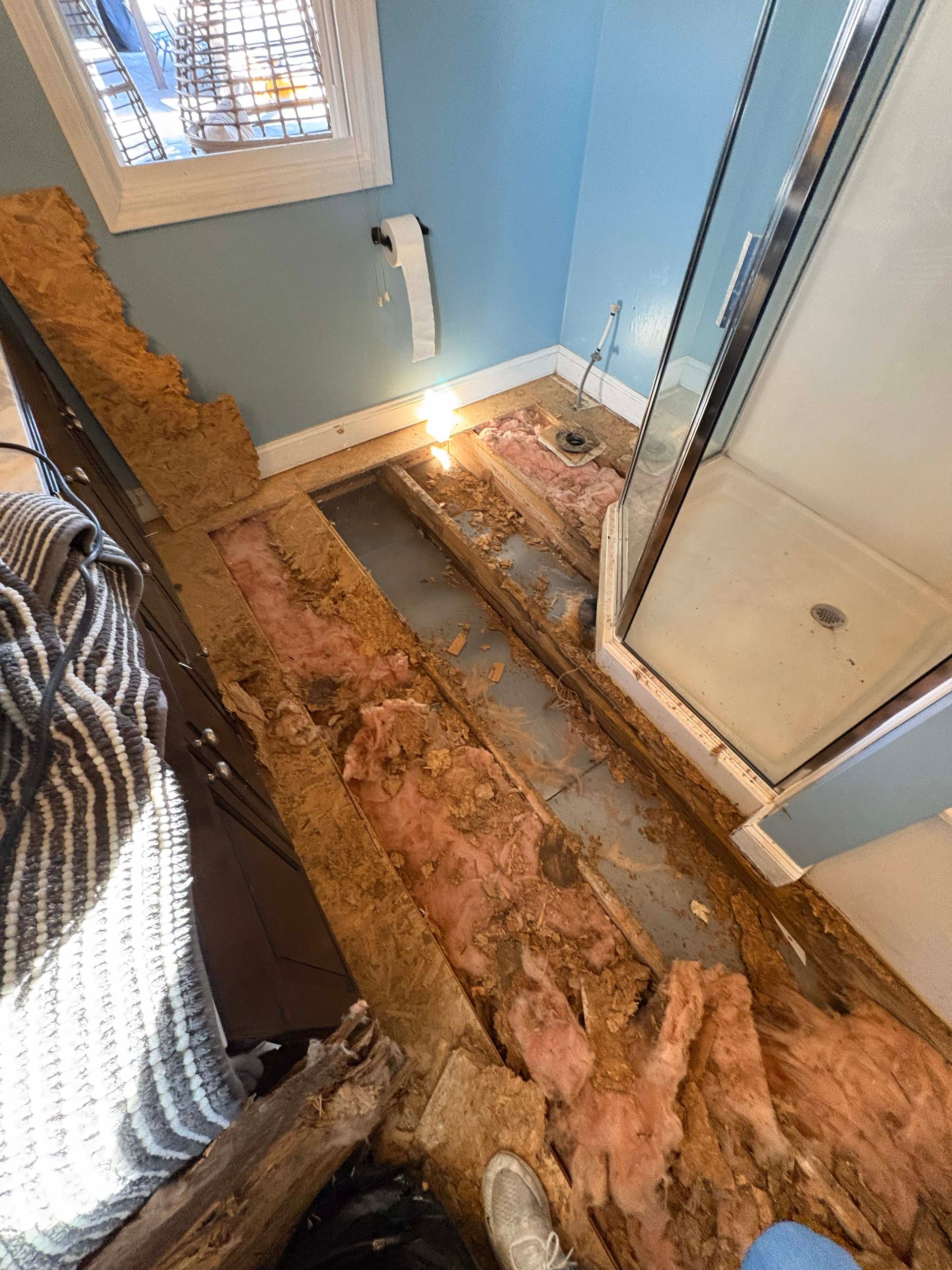 A bathroom floor with plywood removed, exposing wooden joists, pink insulation, and a shower stall in a blue-walled room.