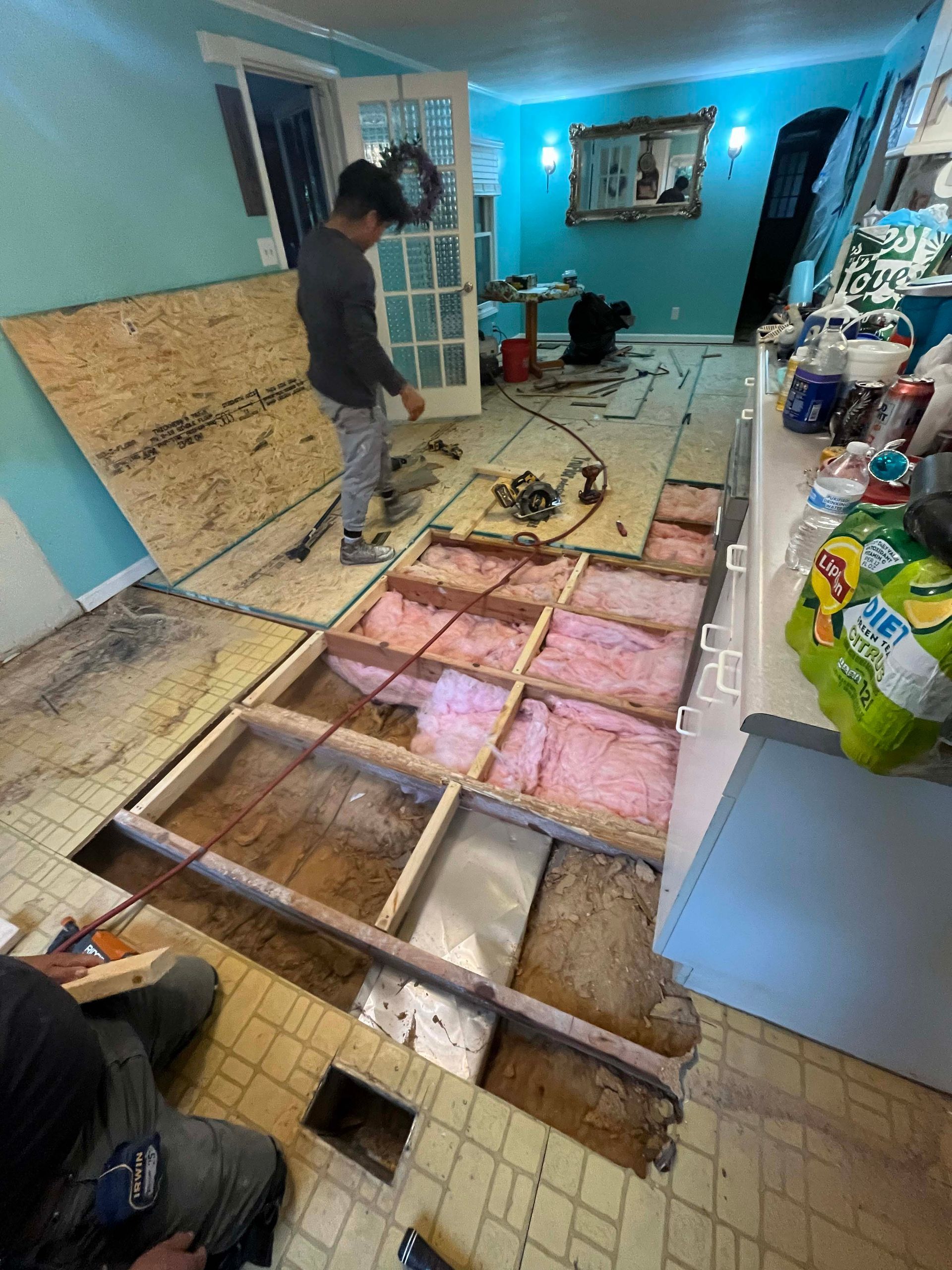 Person inspecting a kitchen floor under renovation, revealing subflooring and insulation. Turquoise walls, open door.