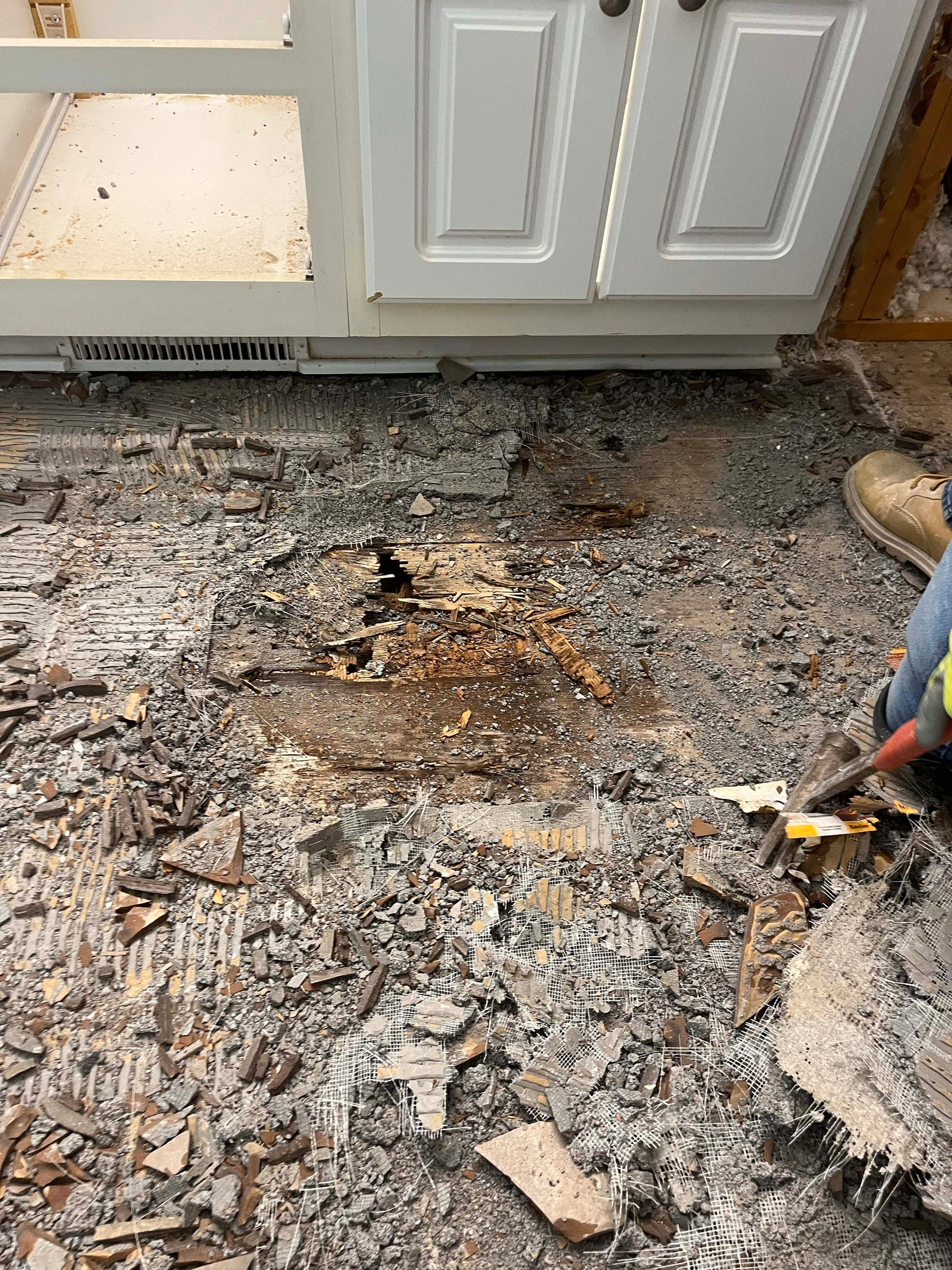 Kitchen floor damaged during renovation; debris scattered in front of a white cabinet.