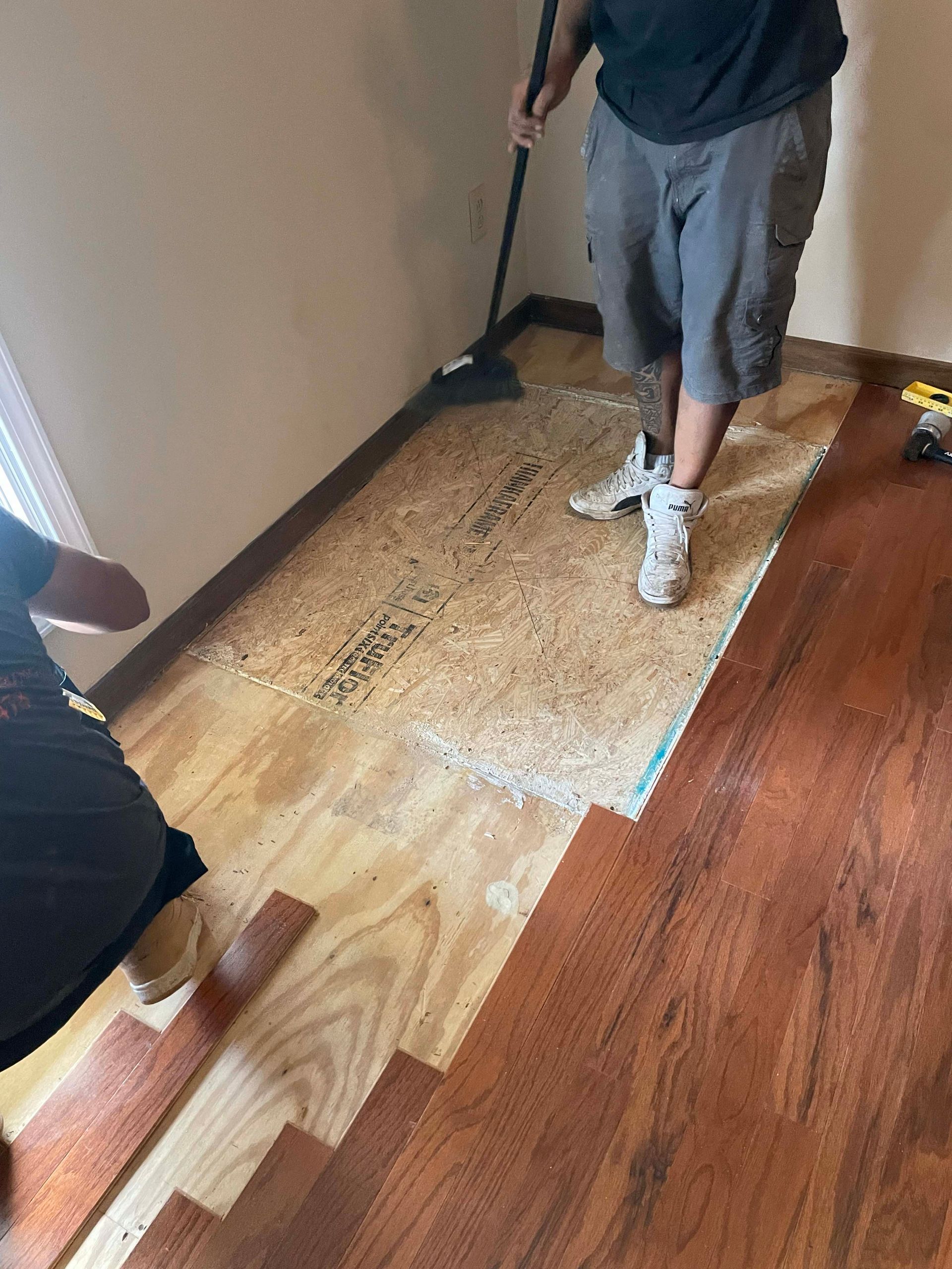 Two people installing wooden floorboards in a room. One sweeps debris, while the other works near a window.