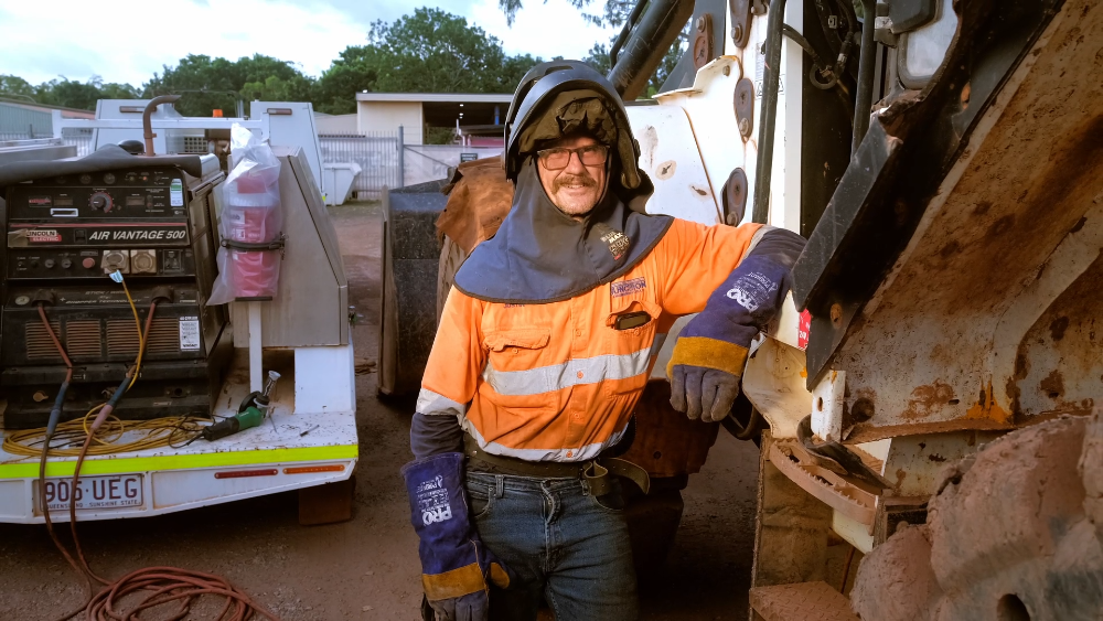 Welder with a helmet and protective gear, leaning on a vehicle. Outdoors, he's smiling, with equipment and buildings in the background — Junction Engineering Group in Evans Landing, QLD