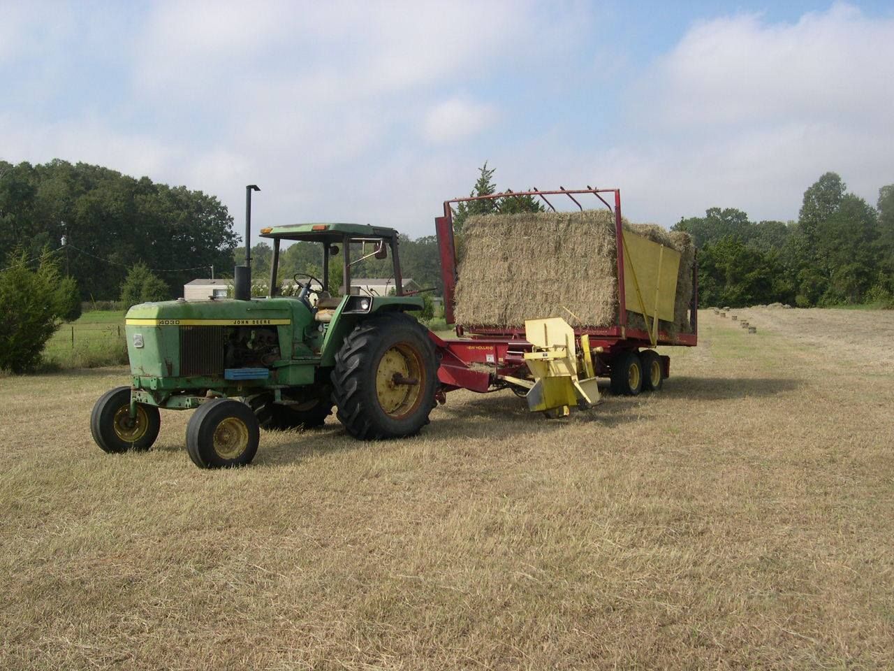Green tractor towing a hay wagon in a dry field under a cloudy sky