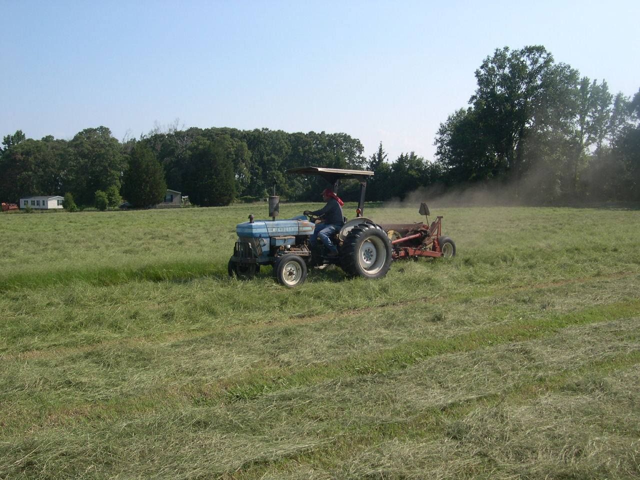 Blue tractor mowing a grassy field, kicking up dust under a clear sky.