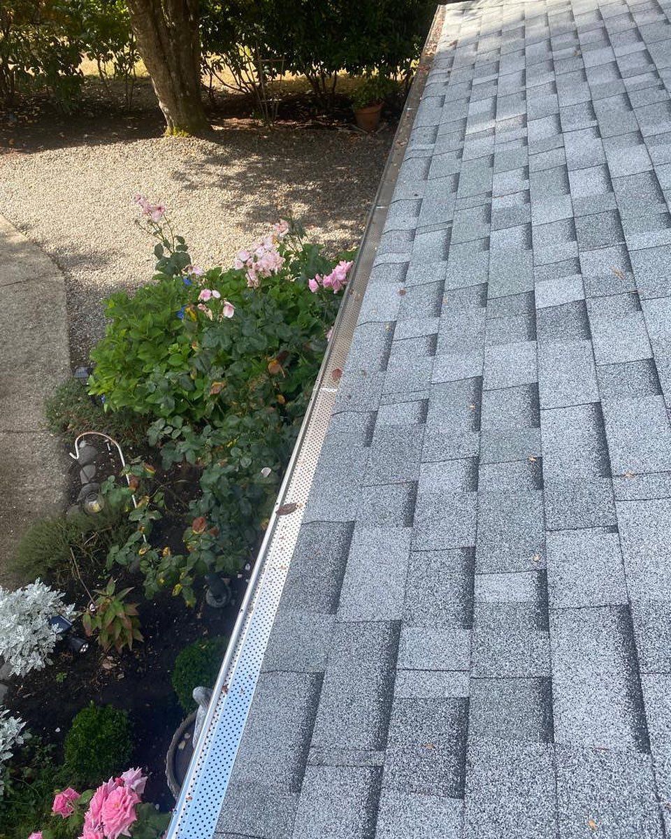 A rooftop with gray shingles and a gutter, beside green plants and pink flowers.