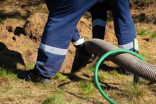 Person in blue work clothes handling a large hose near a hole in the ground.