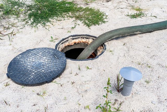Septic tank being pumped out, with a hose inserted, and lid off in sandy ground.