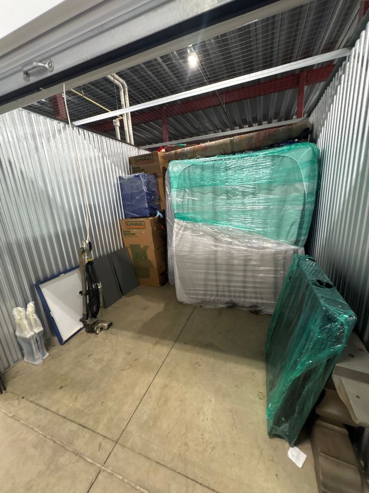 Small storage unit with stacked, plastic-wrapped furniture and boxes along corrugated metal walls