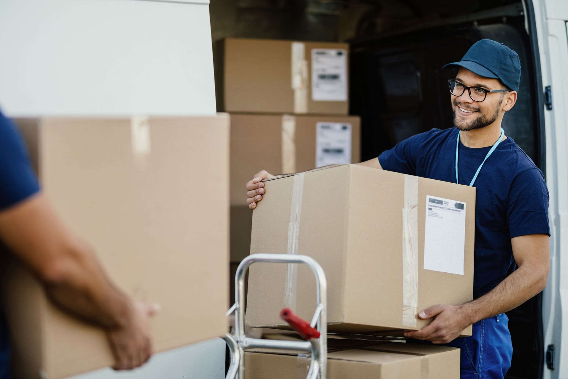 Man carrying a cardboard box in a warehouse, smiling behind a hand truck with more boxes nearby
