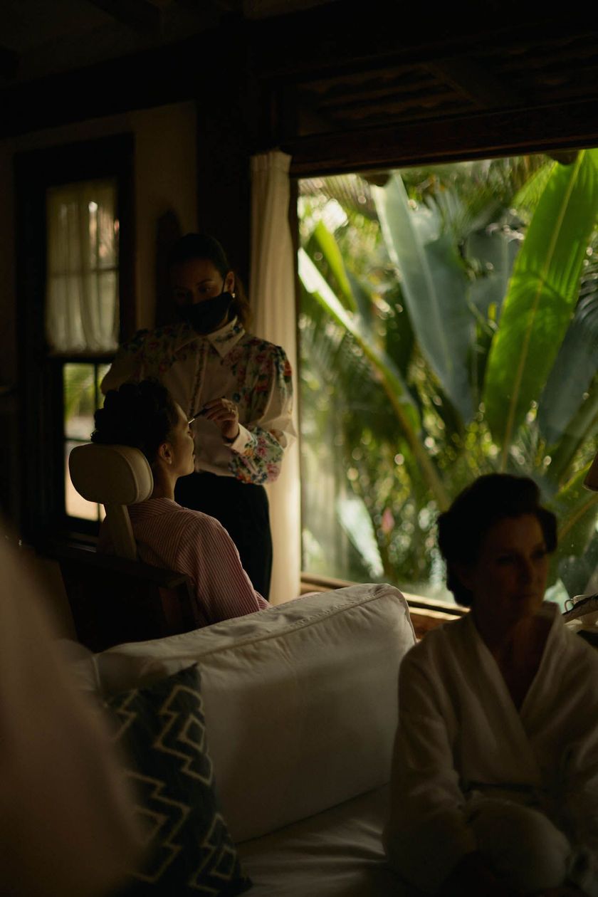 Pessoas reunidas em uma sala de estar pouco iluminada, com a luz do sol e plantas verdejantes visíveis através de uma janela aberta.