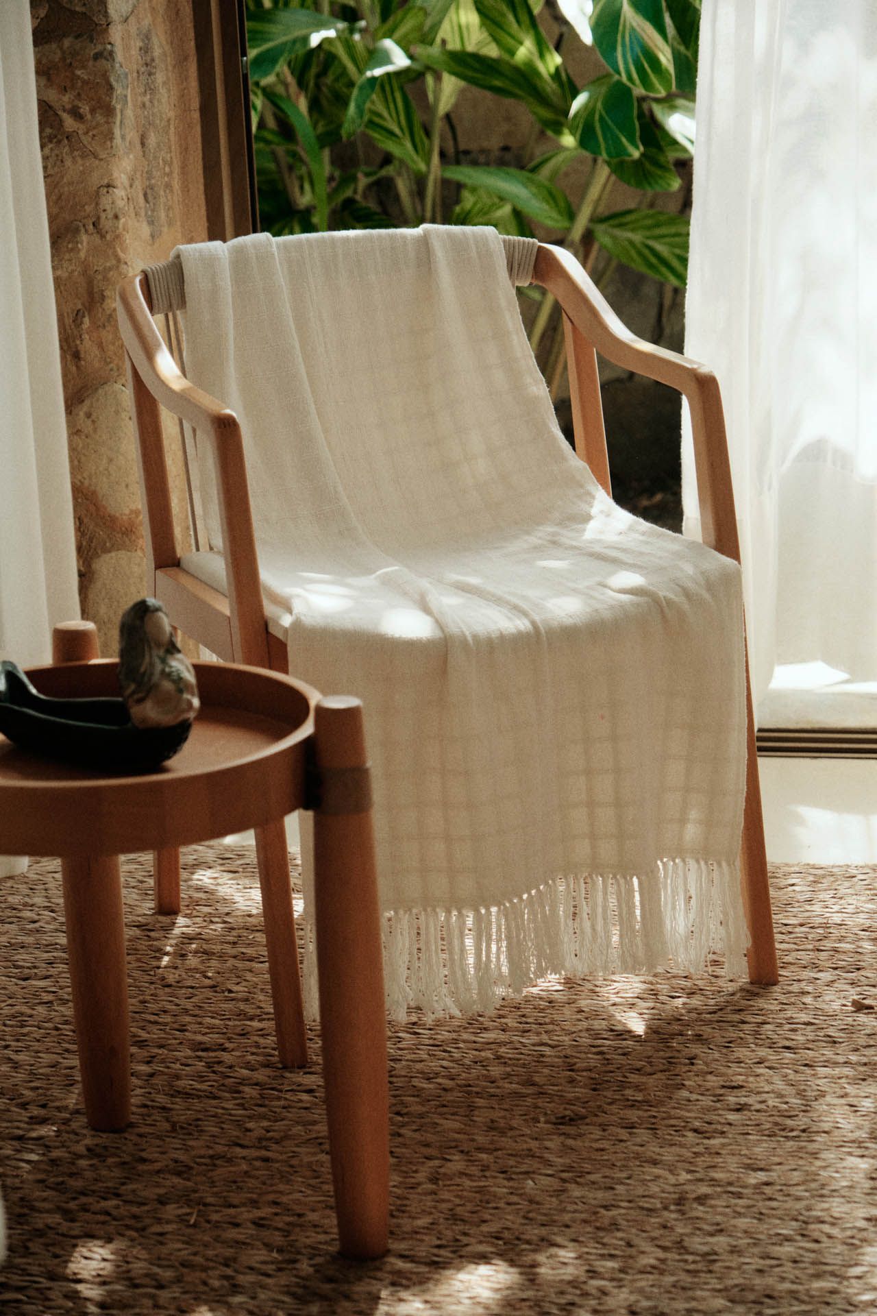 Cadeira de madeira coberta com uma manta creme ao lado de uma mesinha em um quarto ensolarado perto de uma janela.