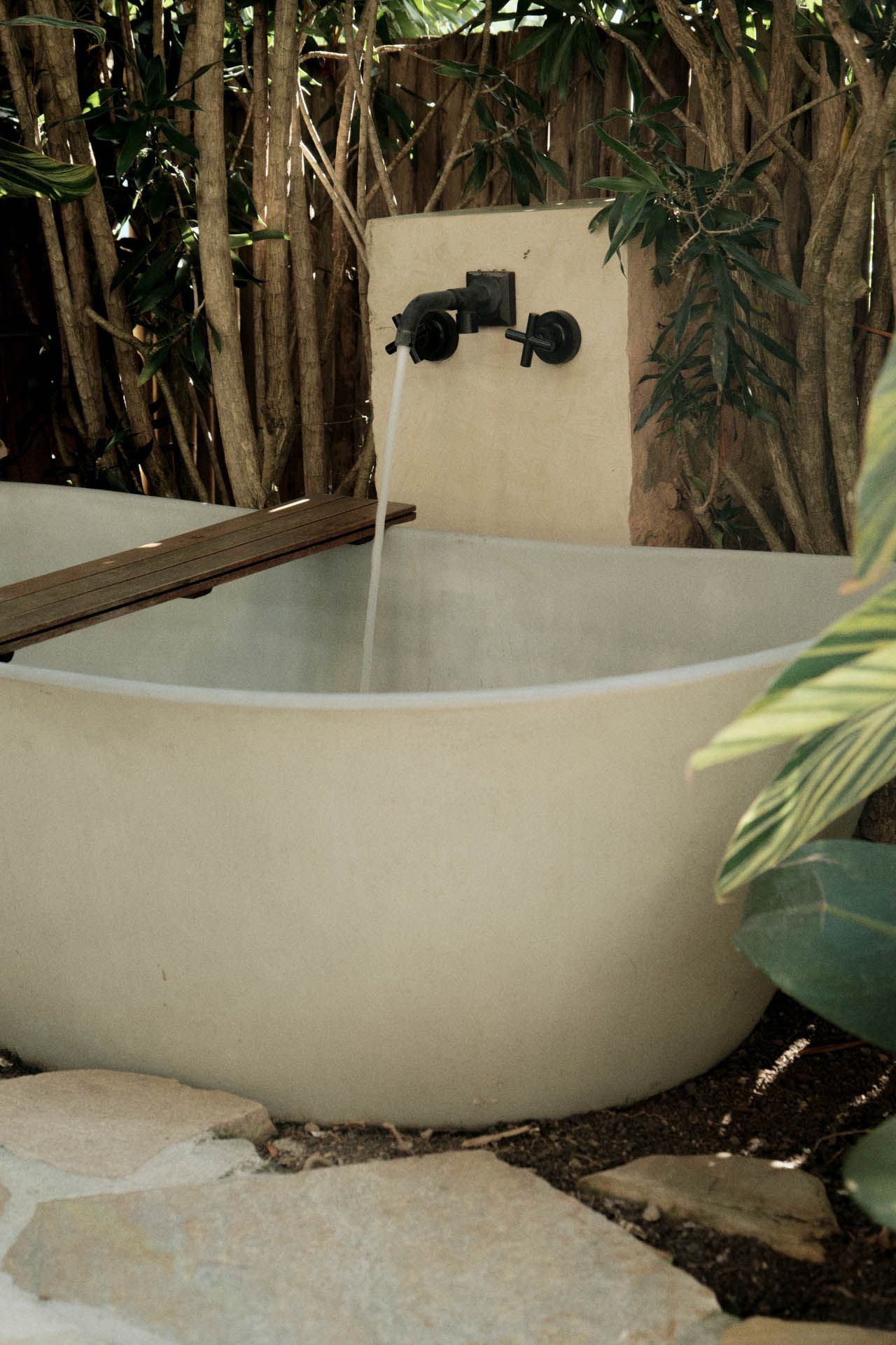 Banheira de pedra moderna para exterior com torneiras pretas fixadas na parede em um jardim tropical.
