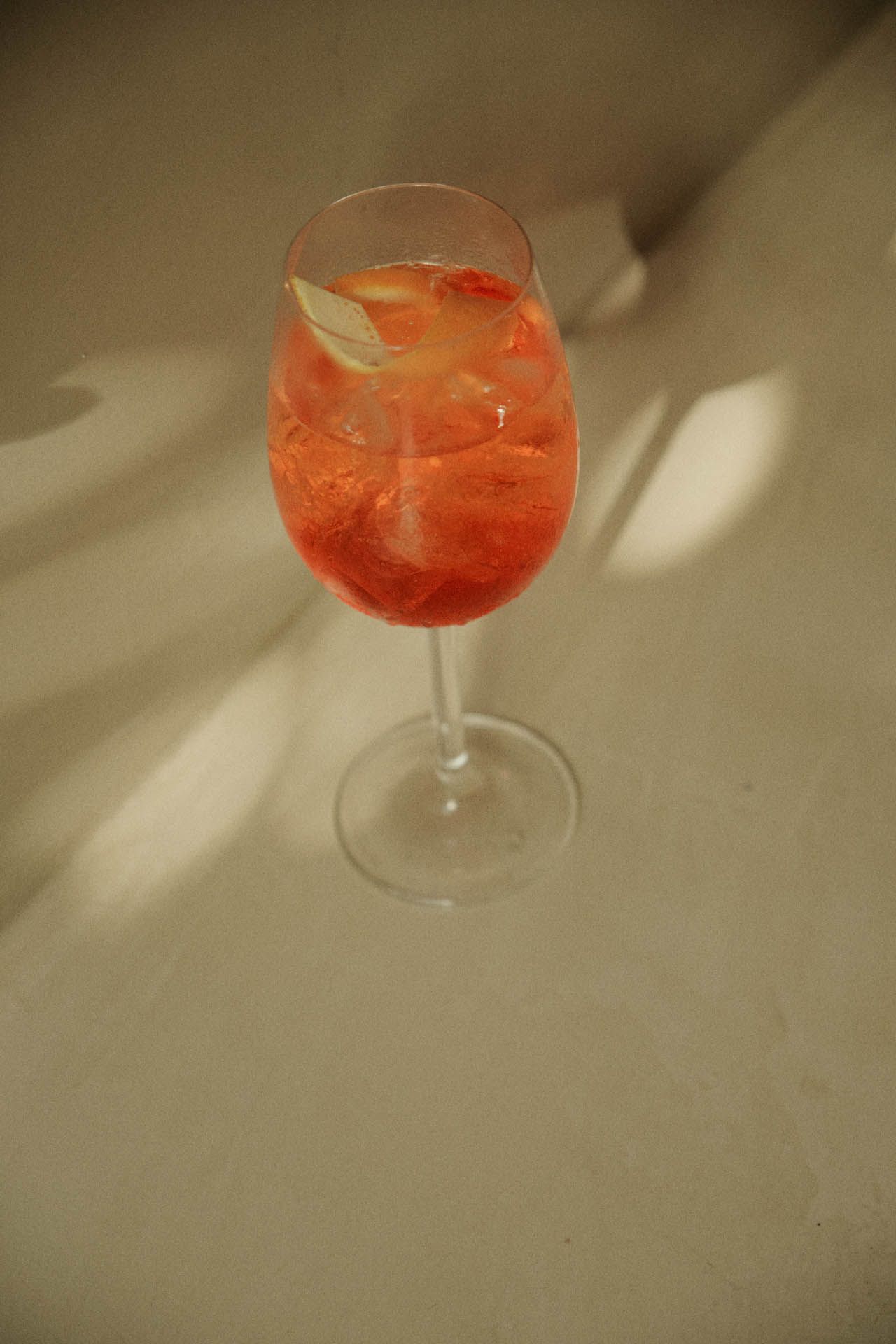 Uma taça de vinho com haste, contendo um coquetel vermelho-alaranjado e gelo sobre uma superfície bege.