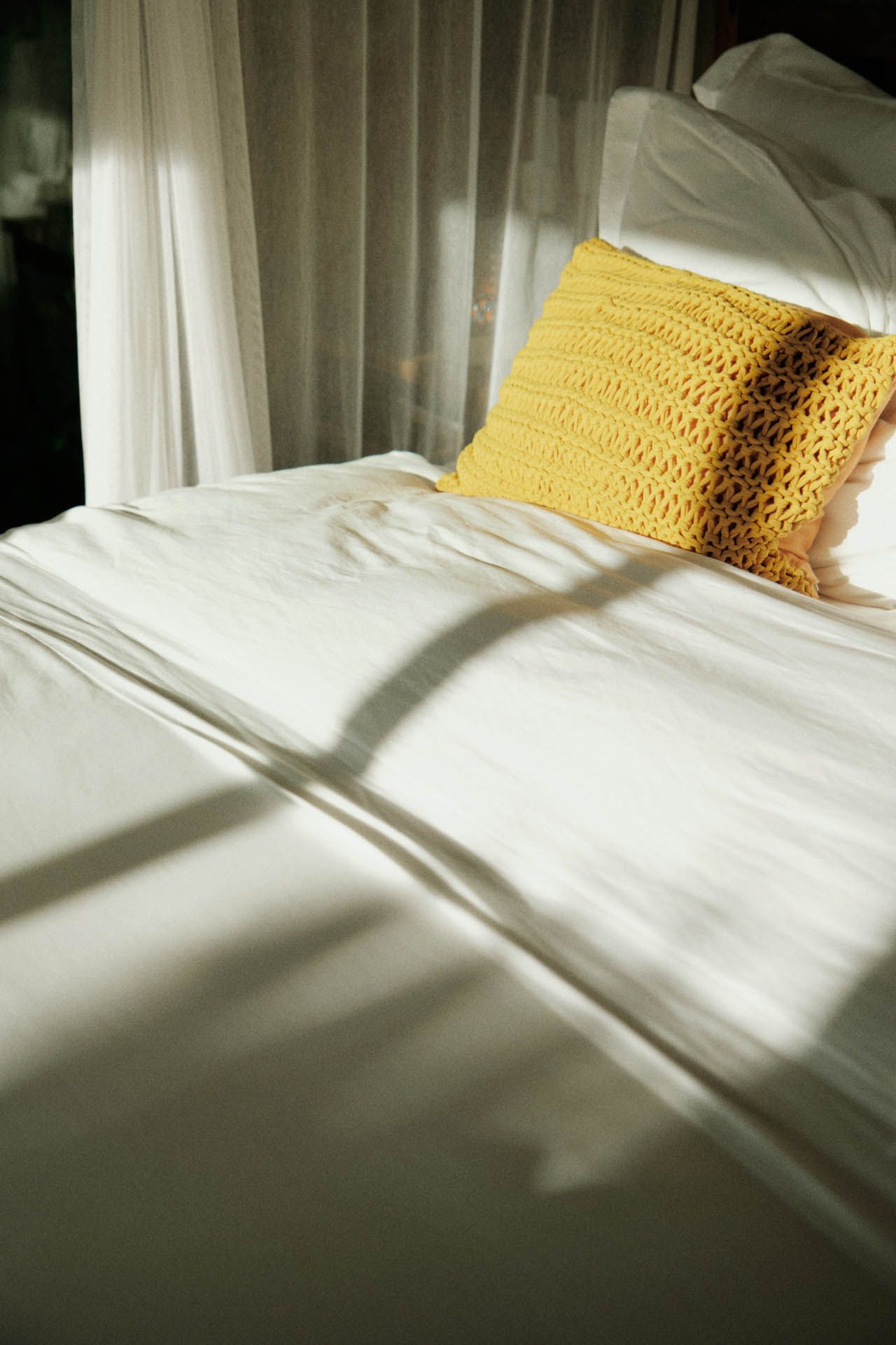 Cama branca banhada de sol com uma almofada amarela texturizada ao lado de cortinas transparentes.