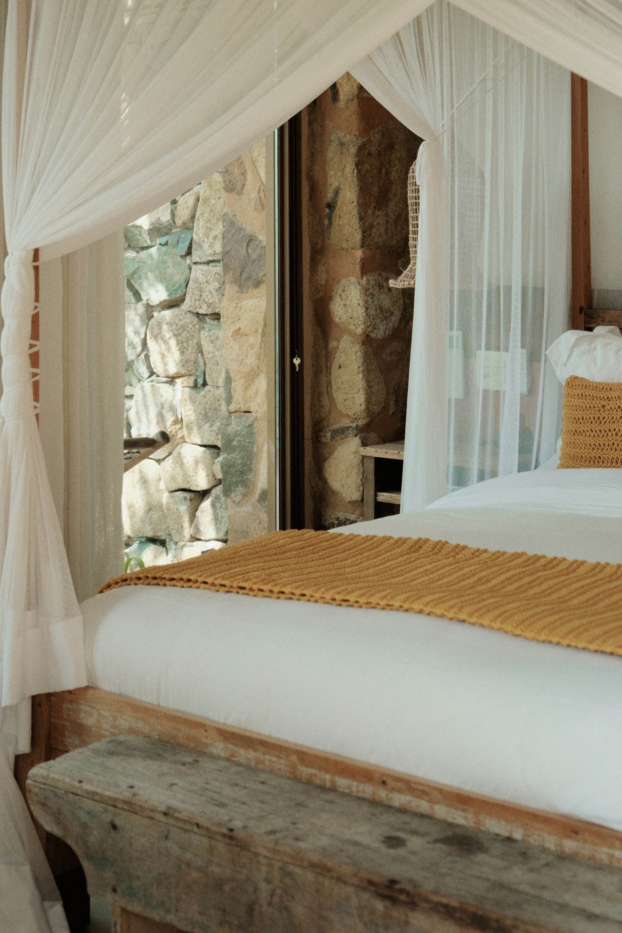 Cama com dossel, cortinas brancas e estrutura de madeira, ao lado de uma parede de pedra e janela.