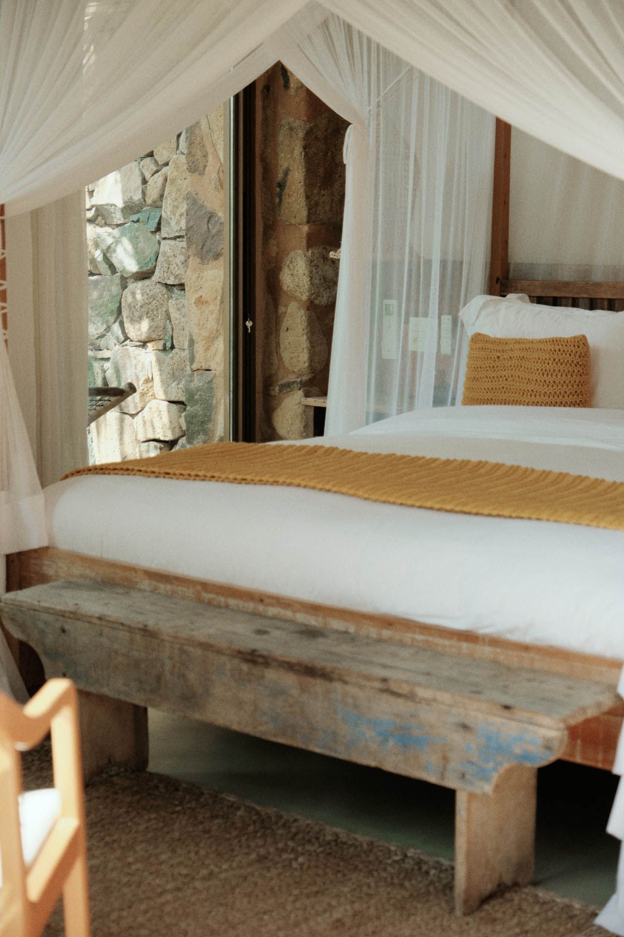 Cama com dossel, cortinas brancas e roupa de cama mostarda em um quarto rústico de pedra.