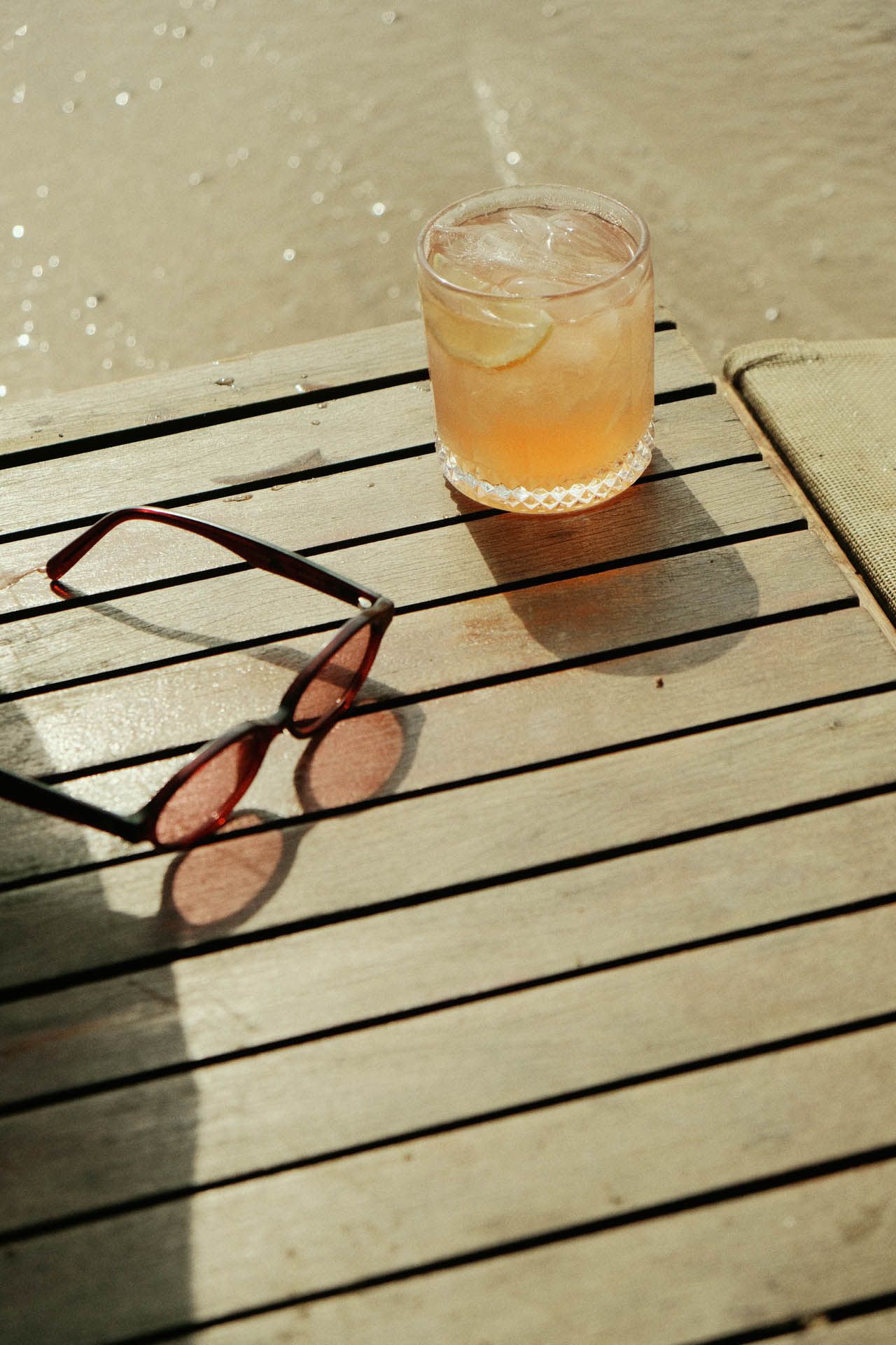Óculos de sol ao lado de um copo de suco de laranja em um deck de madeira ensolarado perto da piscina.