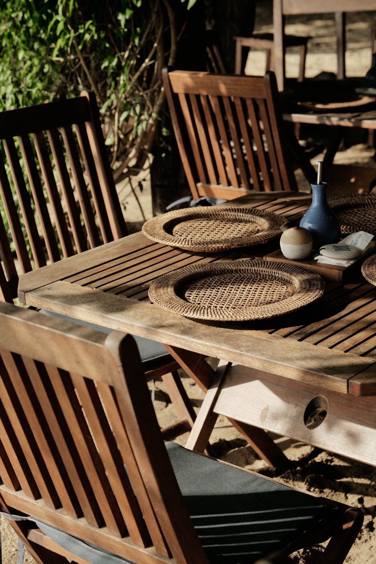 Mesa de pátio de madeira com jogos americanos de vime e vasos de cerâmica em um ambiente externo ensolarado.