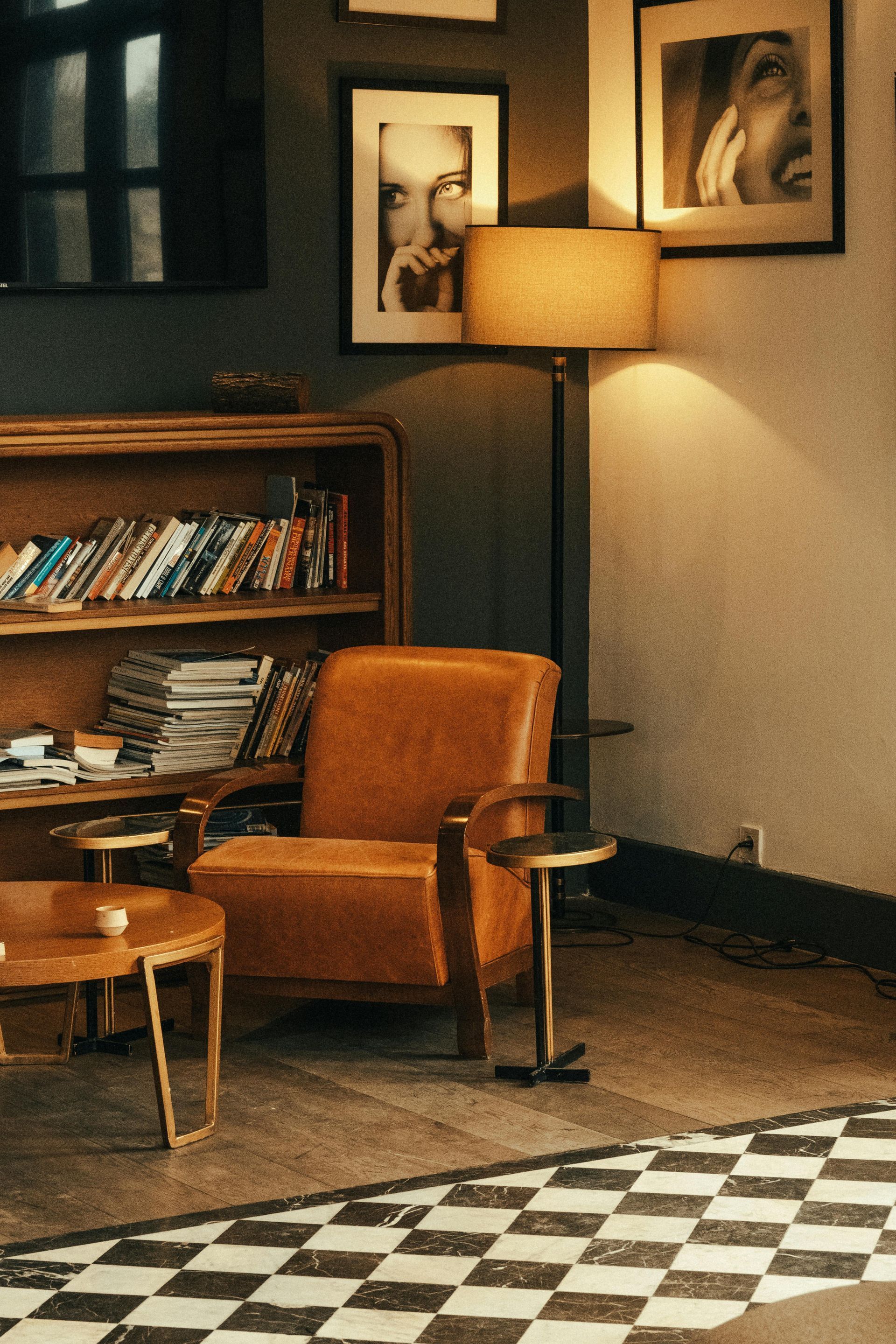 Cozy reading nook with a leather armchair, lamp, and framed art. Bookshelf and checkered floor.
