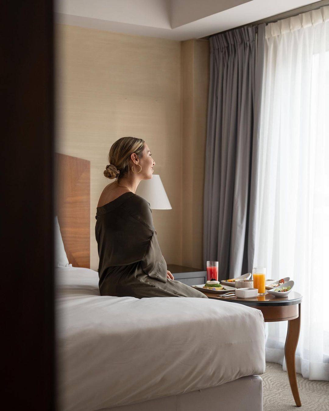 Una mujer está sentada en una cama en una habitación de hotel mirando por la ventana.