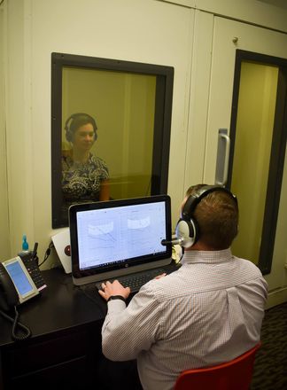 A person using a computer while another sits in a soundproof booth; both wearing headphones.