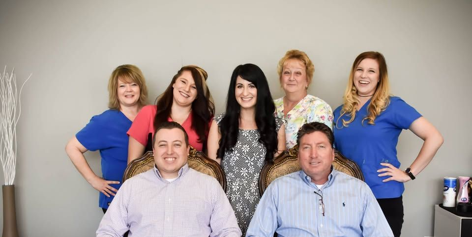Seven people pose in front of a gray wall. Two men sit in ornate chairs. Five women stand behind, smiling.