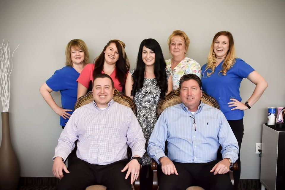 Team photo: Two men seated, six women standing, smiling, against a gray wall.