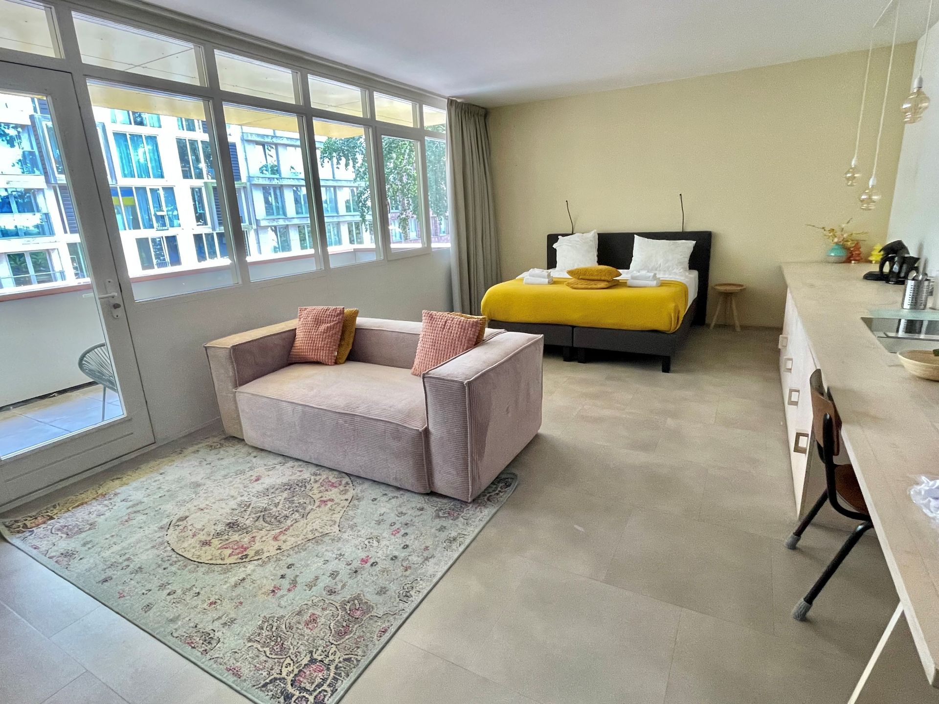 Een lichte hotelkamer is voorzien van een bed met geel beddengoed, een roze bank een bureau bij grote ramen.
