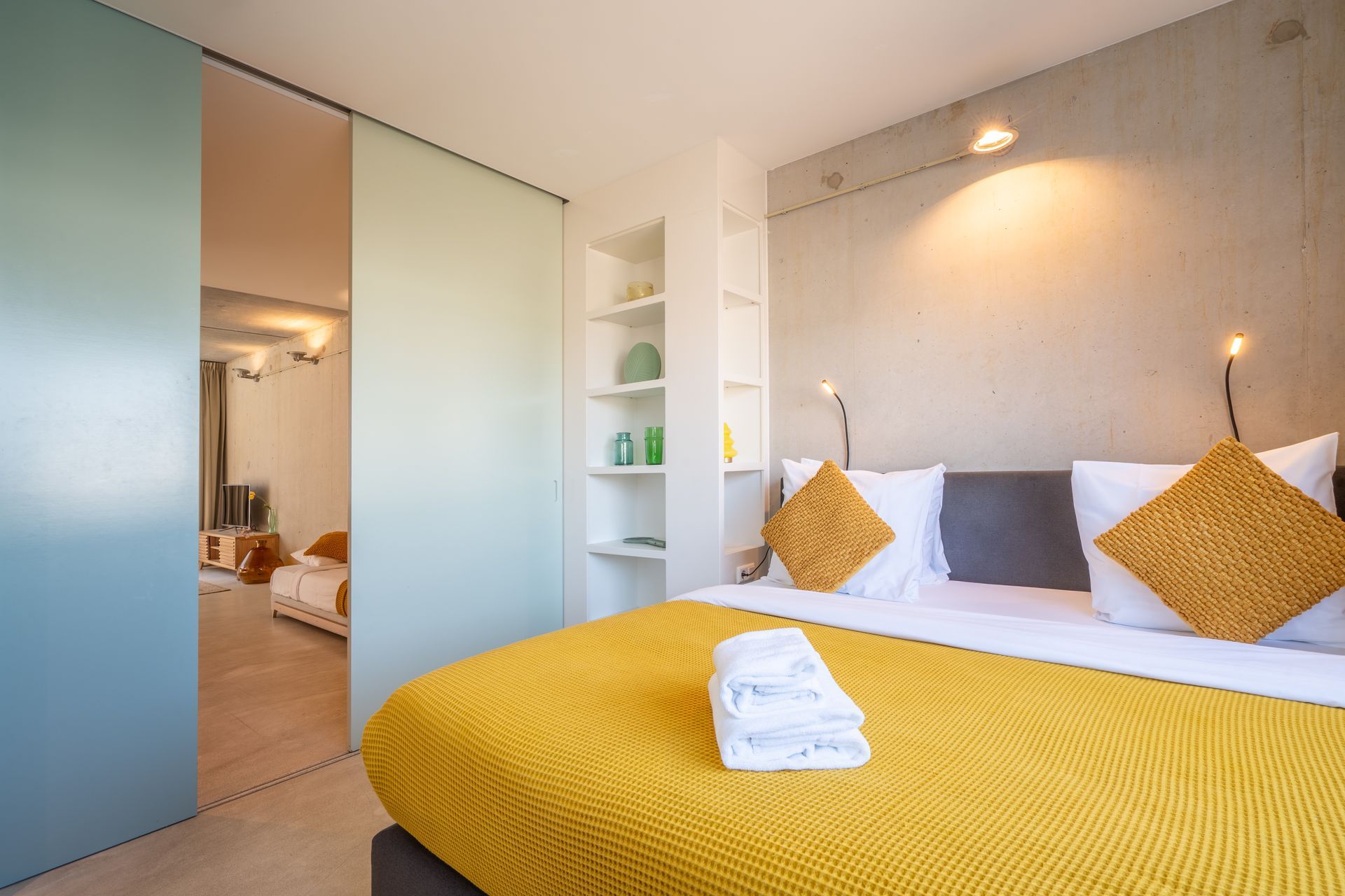 Een moderne slaapkamer met een gele deken en sierkussens op het bed, naast witte planken en een blauwe schuifdeur.