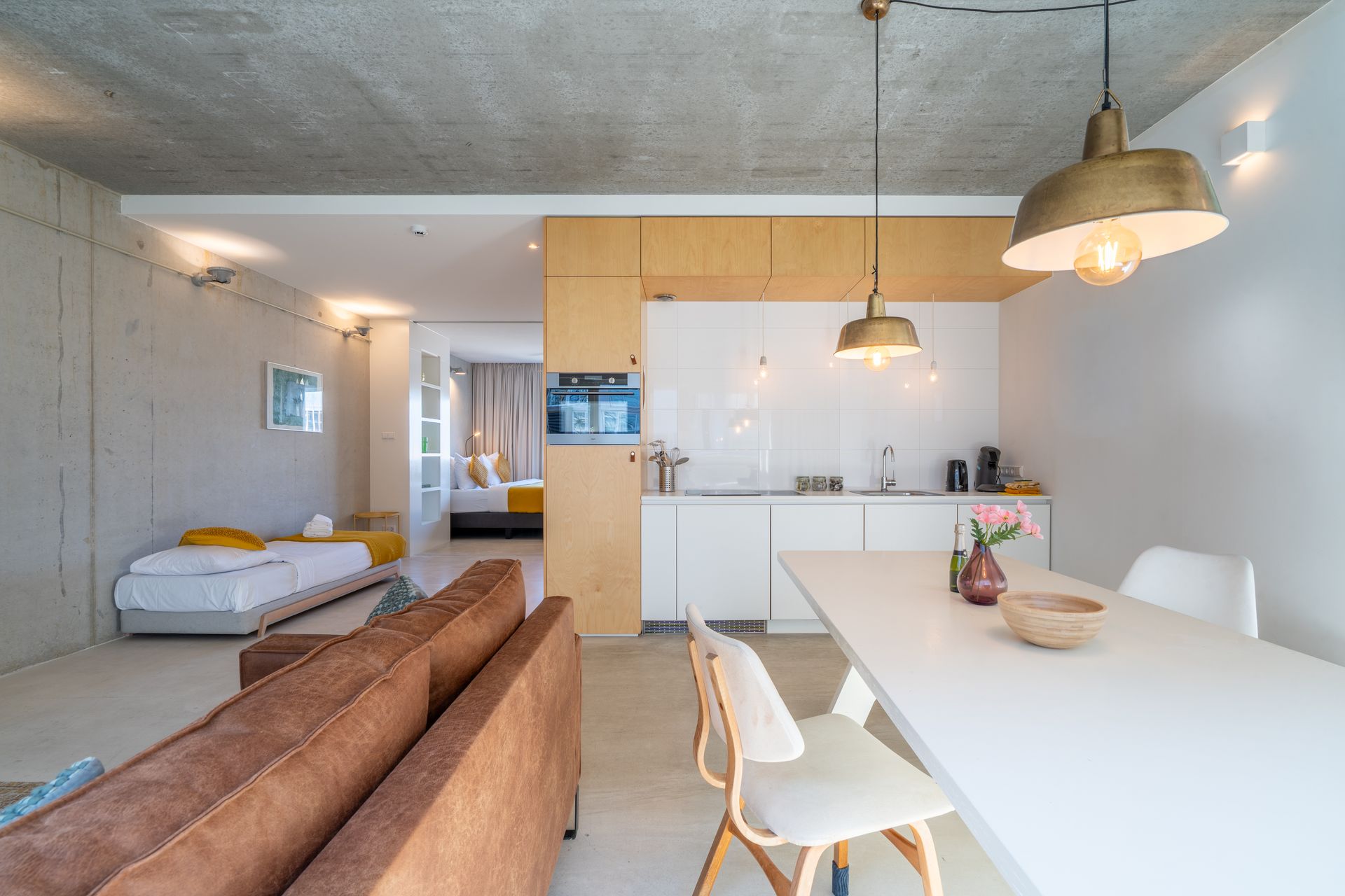 Een modern open appartement met een keuken, eethoek, bruine bank en een zichtbare slaapkamer op de achtergrond.