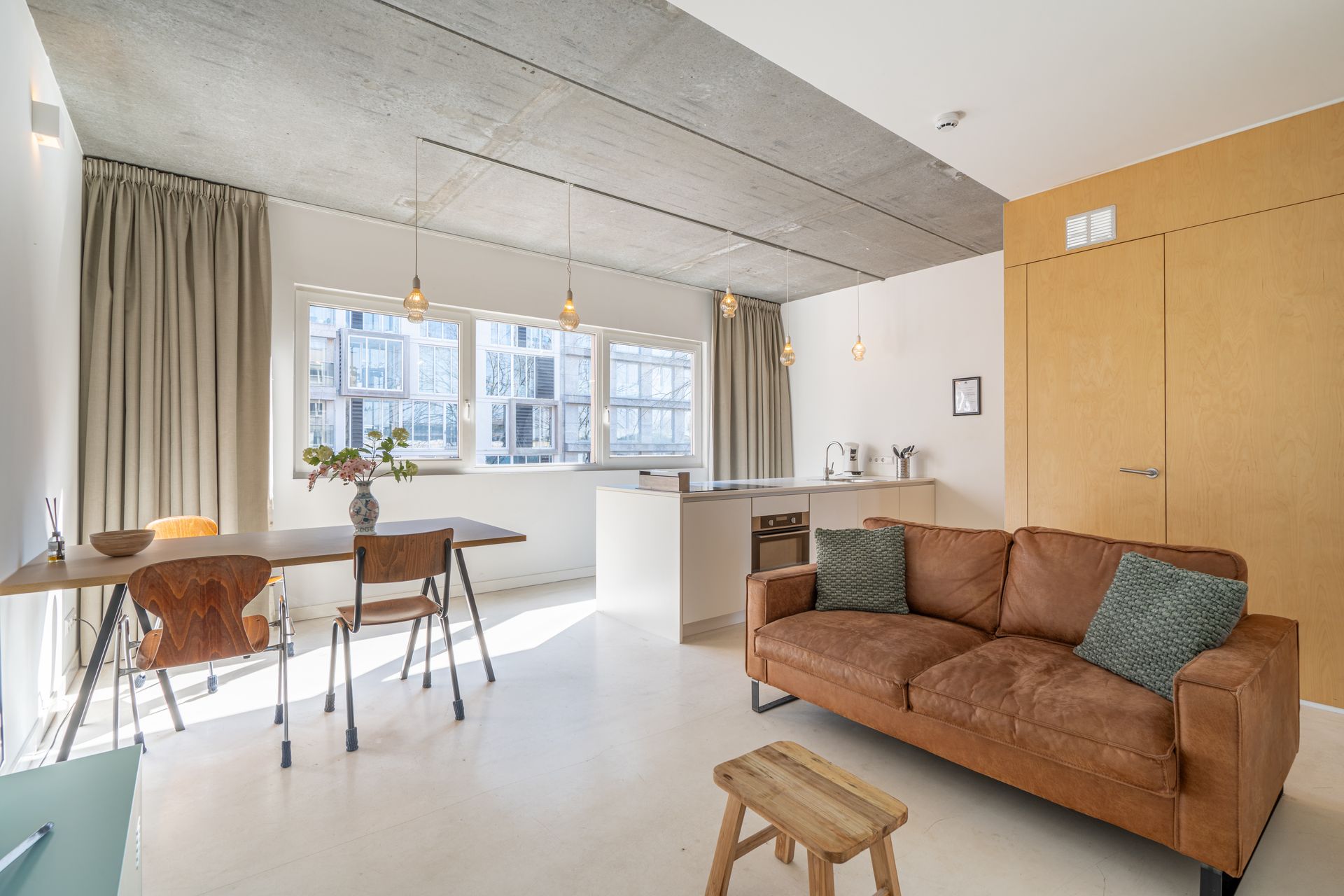 Modern appartement met betonnen plafond, bruine leren bank, eettafel en lichte houten kasten in een open ruimte.