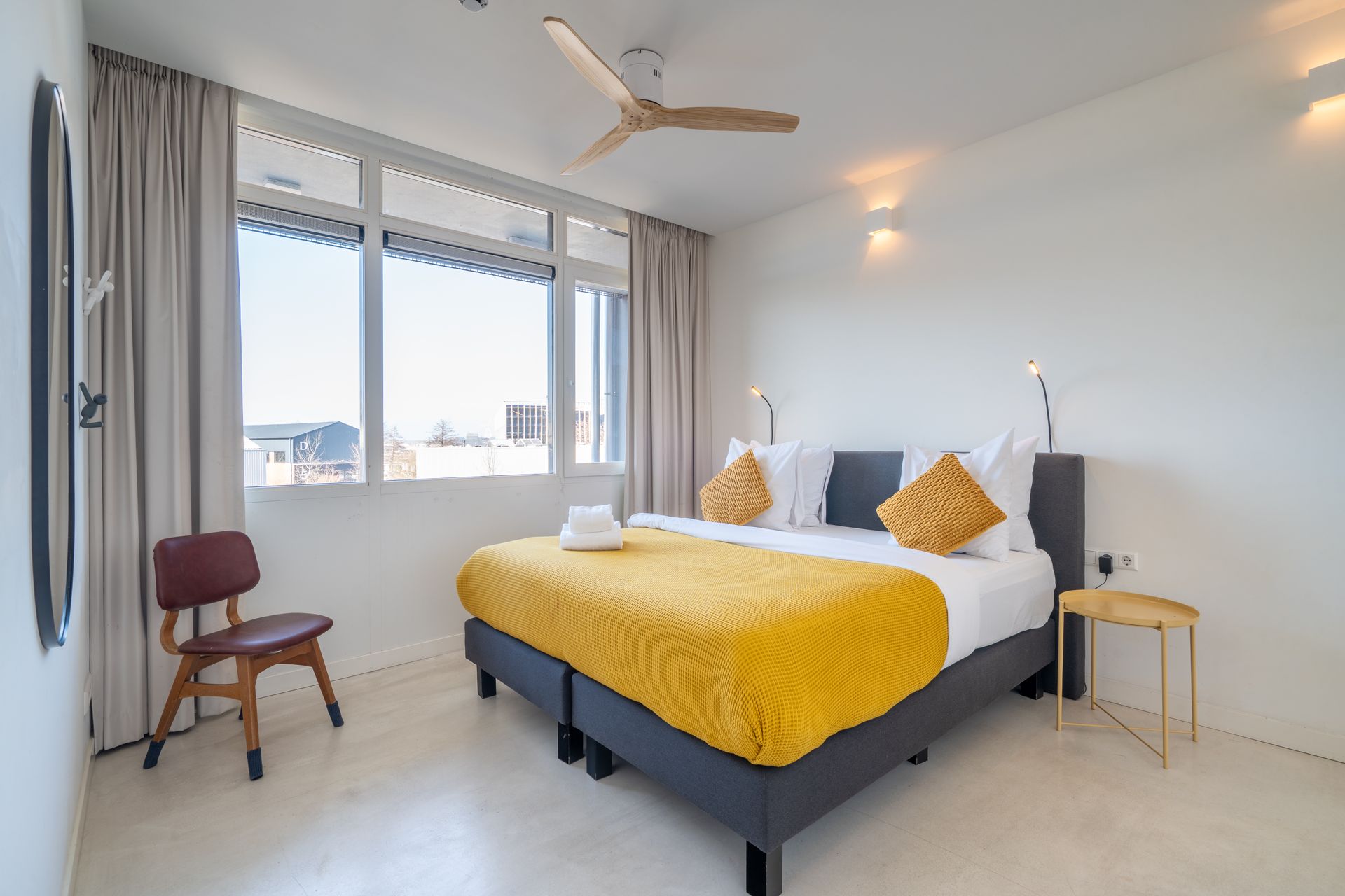 Een lichte slaapkamer met een tweepersoonsbed (queensize, grote ramen met beige gordijnen.