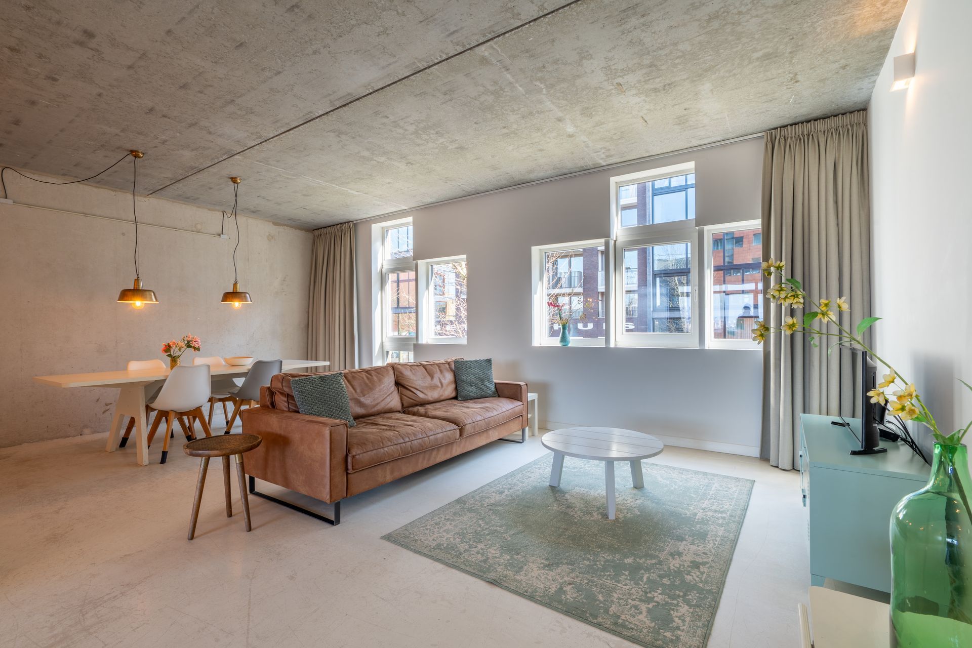 Moderne woonkamer met een leren bank, eethoek, betonnen plafond en grote ramen met lichtgekleurde gordijnen.