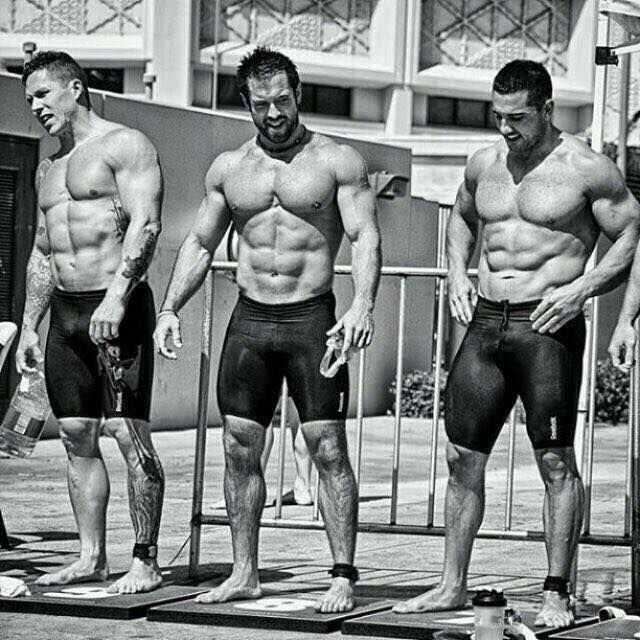 Three shirtless bodybuilders standing on a stage, posing in black trunks under bright lights.