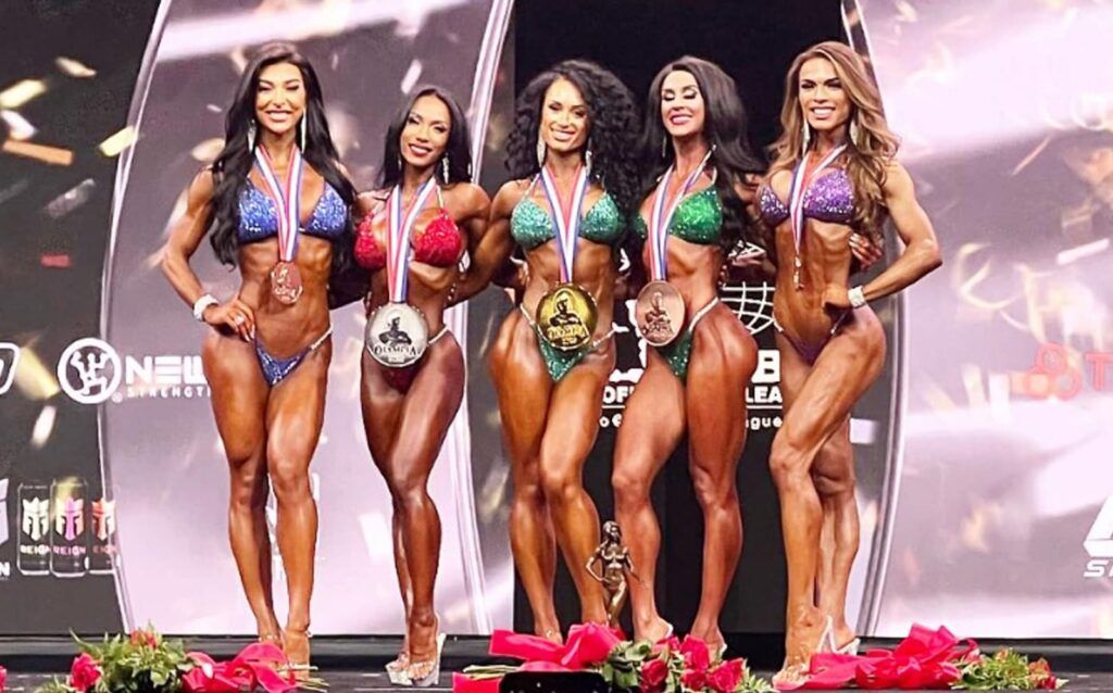 Five female bodybuilders in colorful bikinis posing with medals on a bodybuilding stage