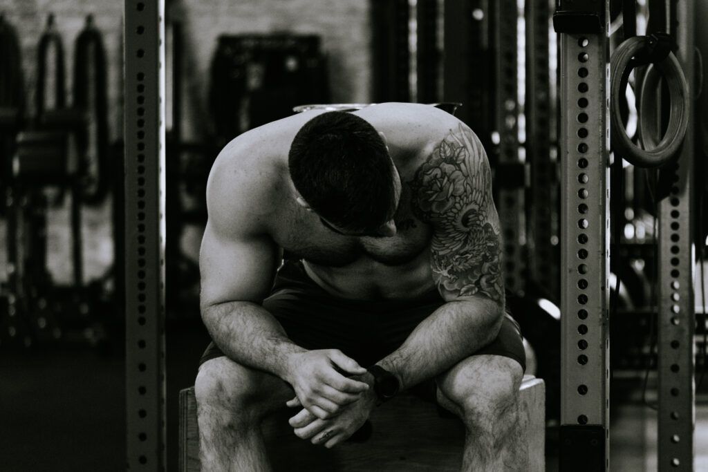 Man sitting on a gym bench, head bowed and hands clasped, in a dim weight room