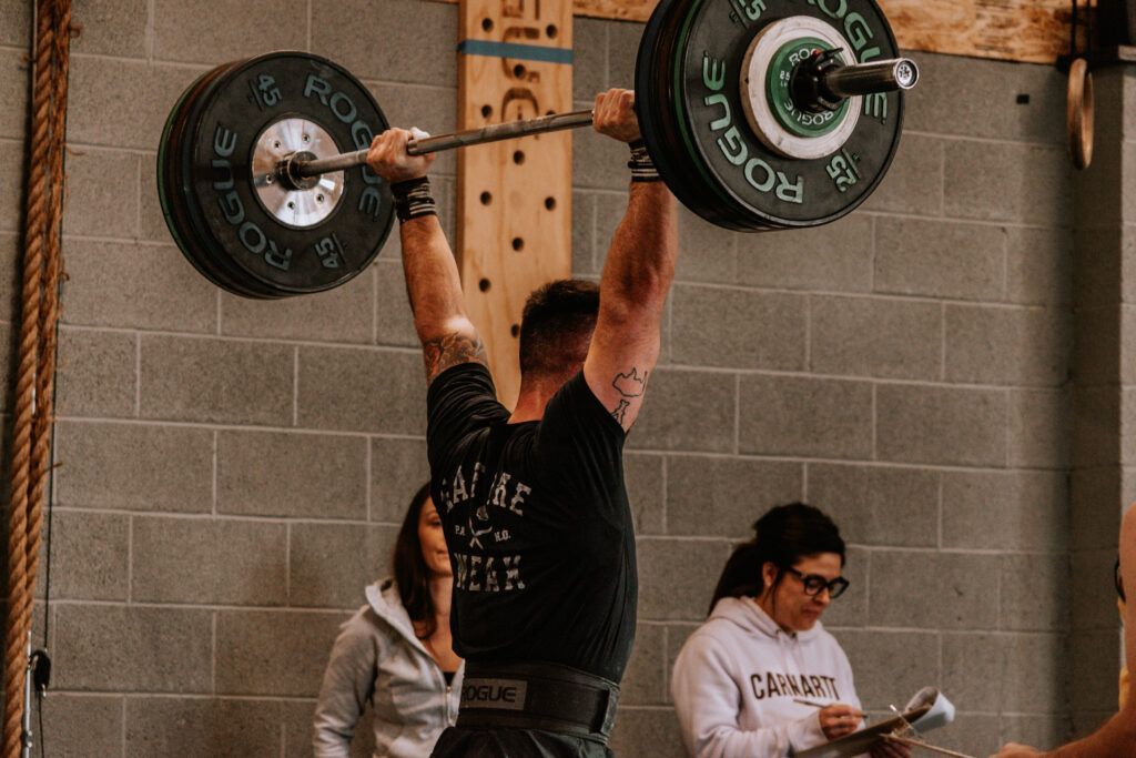 Weightlifter holding a barbell overhead in a gym, with two people watching nearby