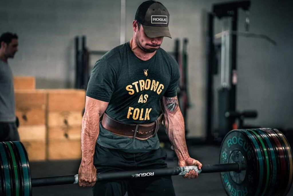 Man deadlifting a heavily loaded barbell in a gym, wearing a shirt that says “STRONG AS FOLK.”