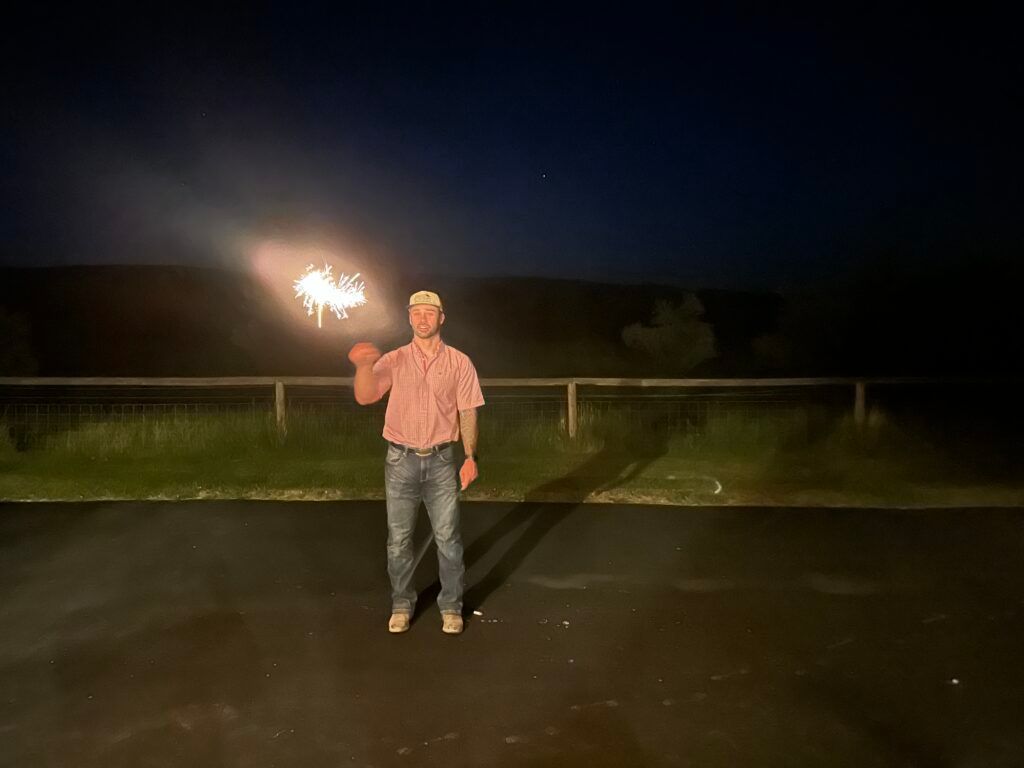 Person standing at night in a parking lot, holding a lit sparkler.