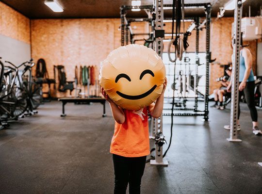 Person holding a large smiley emoji face in a gym with exercise equipment in the background