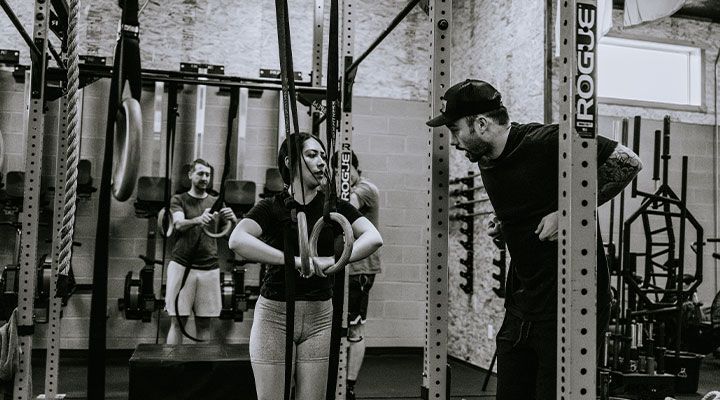 Three people lifting weights in a gym, with one coach spotting them between racks.