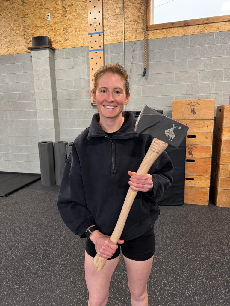 Smiling woman in a gym holding a sledgehammer with wood boxes and cinder blocks behind her