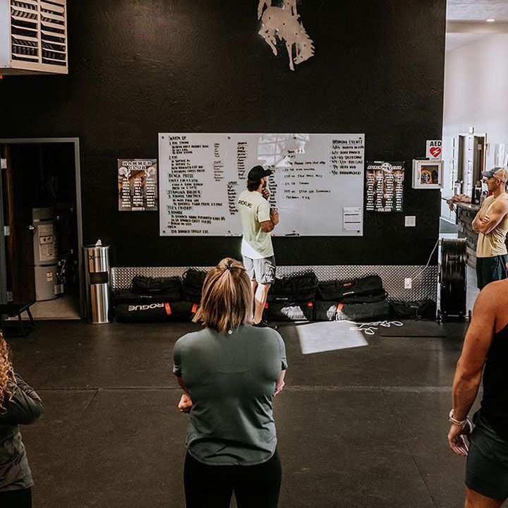 Person speaking in a gym while others listen, facing a large whiteboard workout plan.