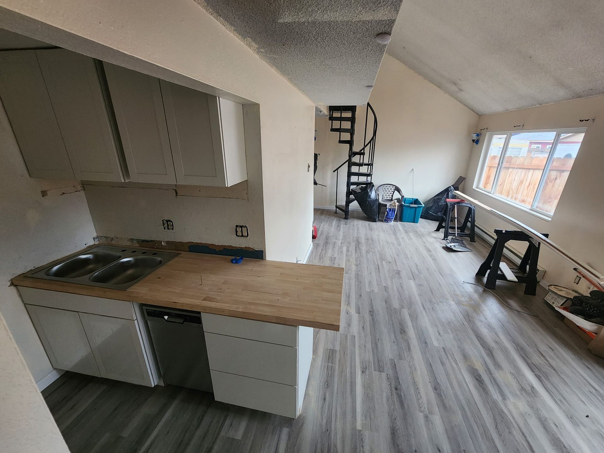 Kitchen with white cabinets, wood countertop, sink, and dishwasher. Gray flooring, spiral staircase, and window.