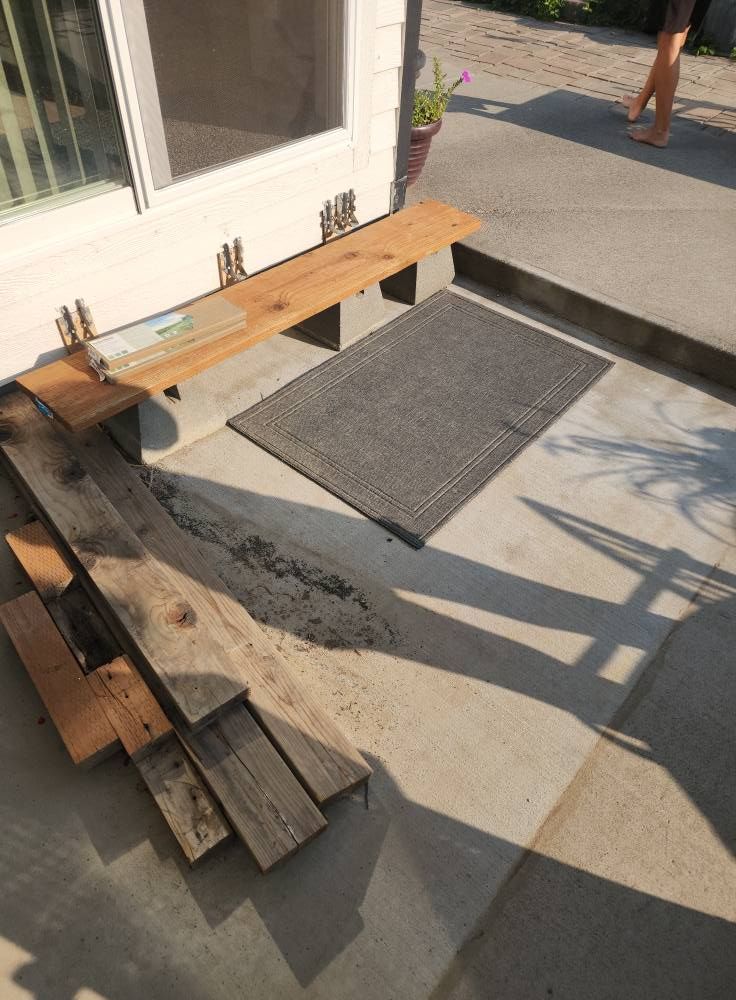 Wooden bench next to a door with a doormat on concrete. Sunlight casts shadows.