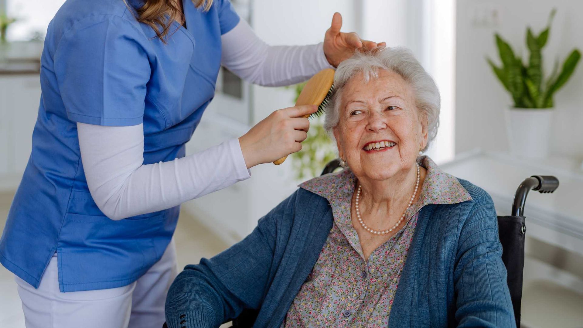 A nurse is brushing the hair of an elderly woman in a wheelchair.