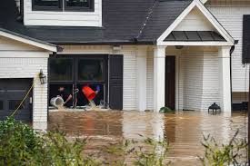House partially flooded; people bailing water from interior with buckets. Brown floodwater surrounds the structure.