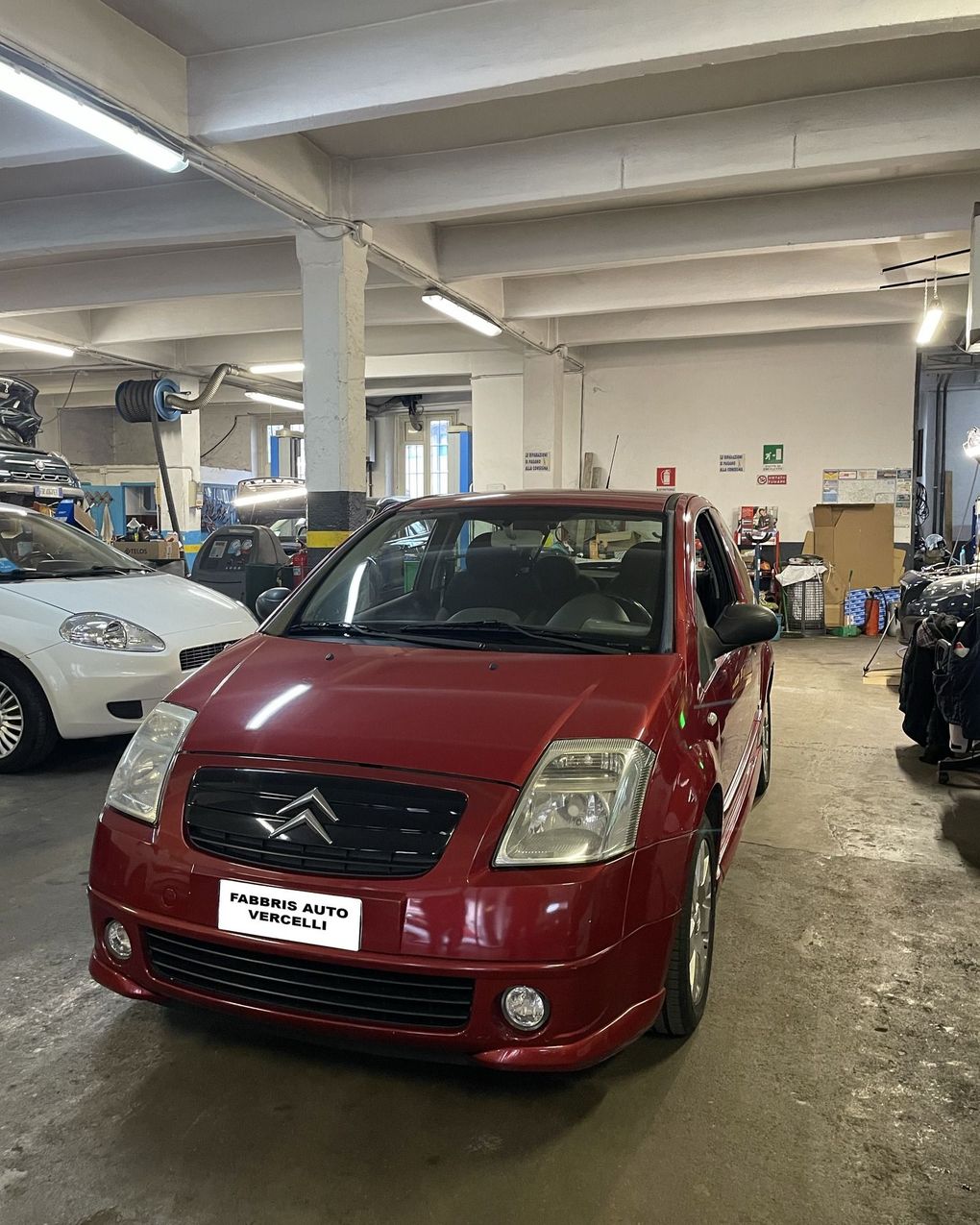 Un'auto Citroën rossa parcheggiata all'interno di un'officina, insieme ad altri veicoli e con le luci accese.