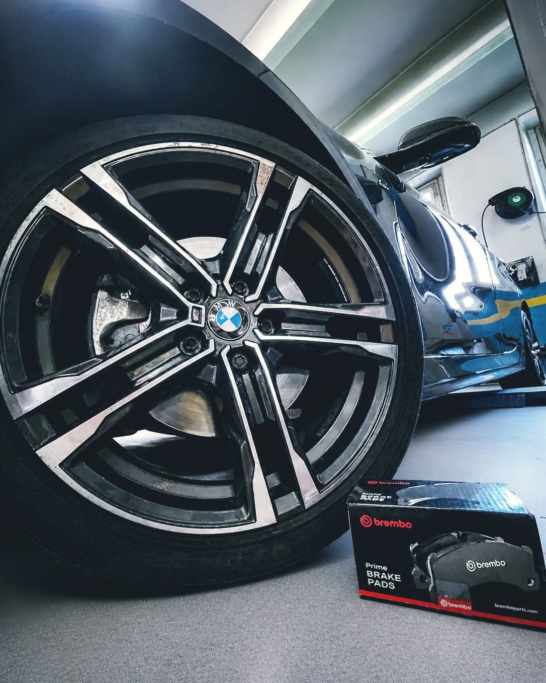 Primo piano di una ruota BMW nera con cerchi argentati in un garage, con una scatola di pastiglie dei freni sul pavimento.