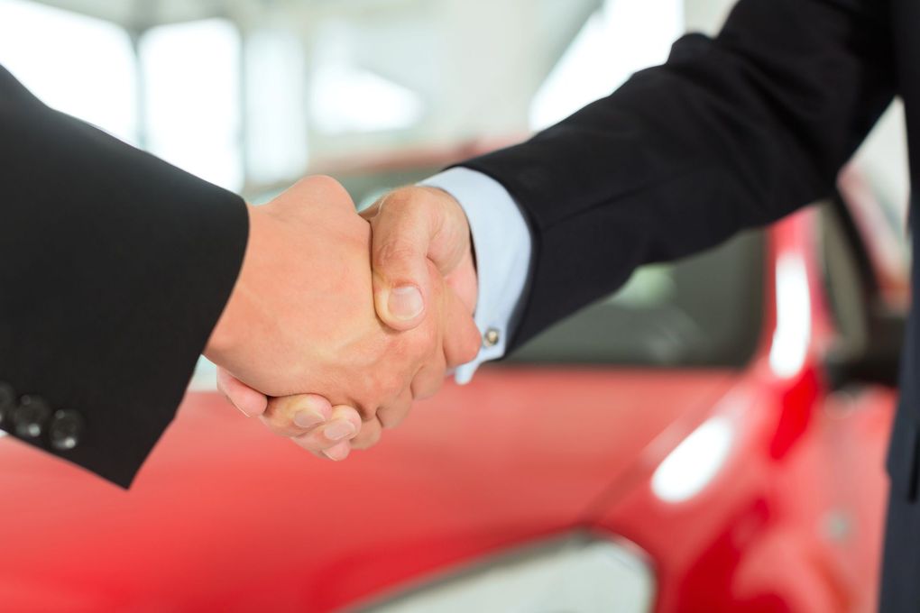 Una stretta di mano davanti a un'auto rossa, a suggerire un accordo commerciale o una vendita.