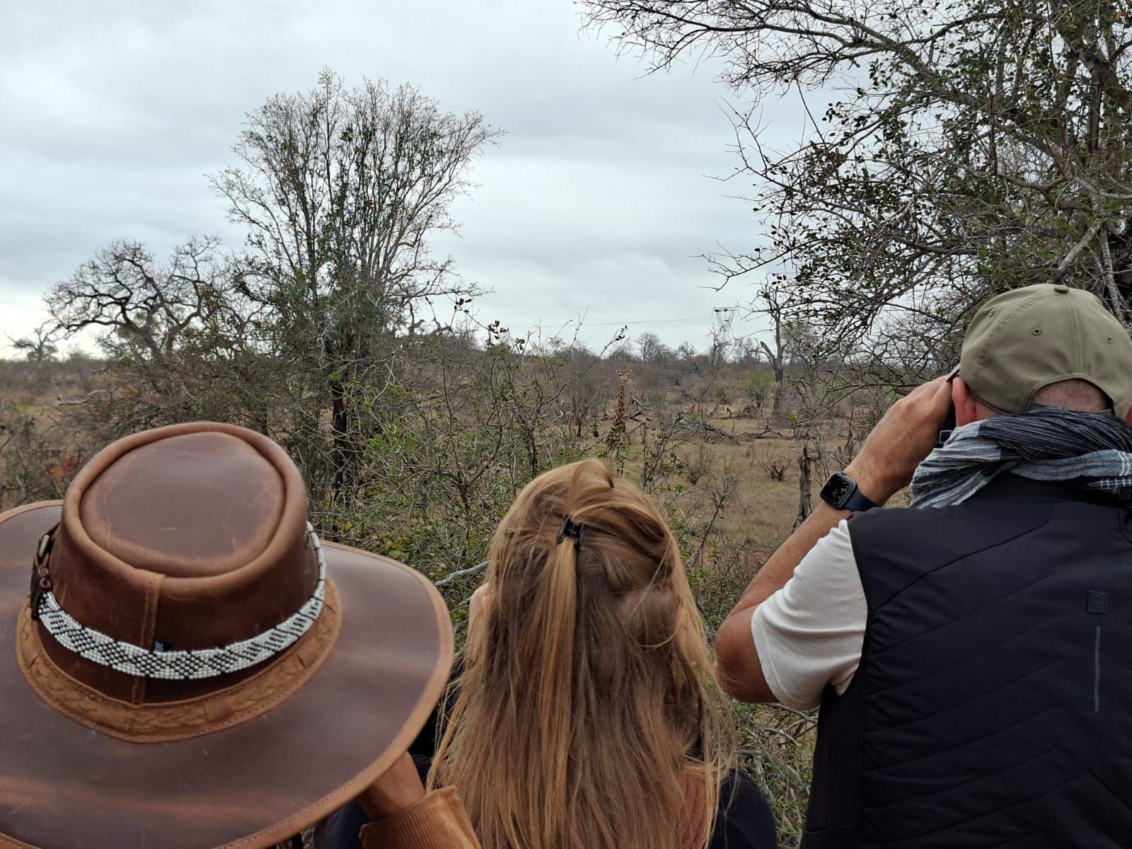 Three people in hats overlook a dry savanna with scattered trees under a cloudy sky.